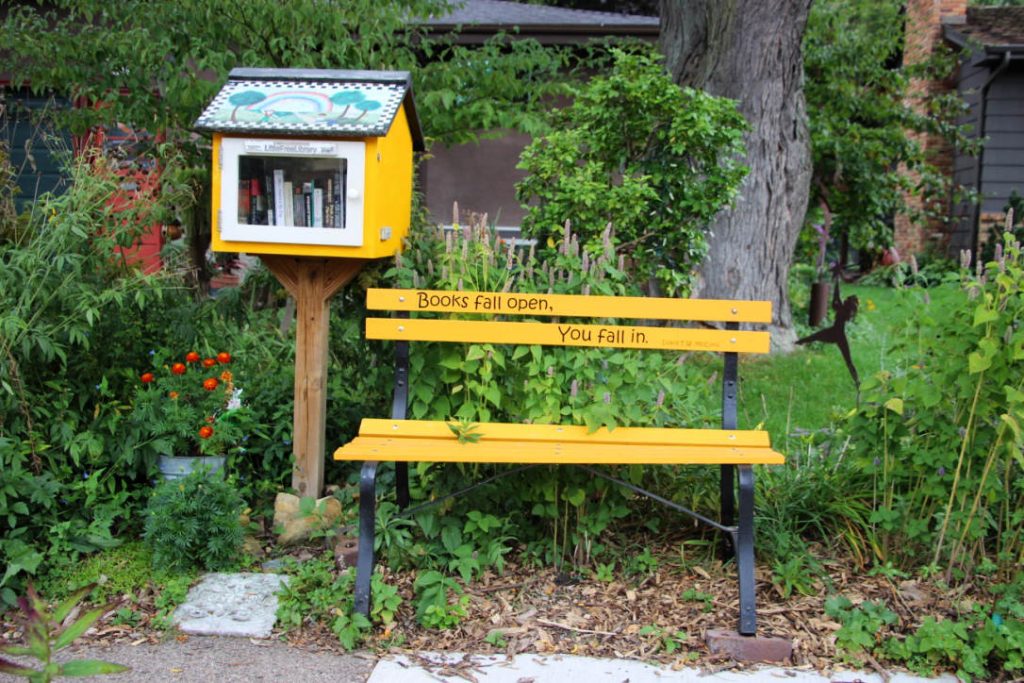 Decorated wooden box of books on a wooden post next to a yellow bench with text reading Books fall open, You fall in.