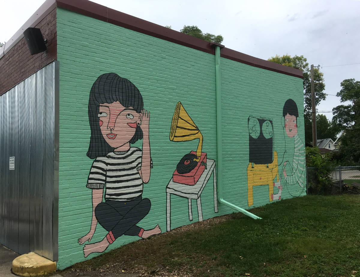 Painting on a green exterior wall of a building with a girl listening to an old victrola record player and a boy with headphones and reel-to-reel tape recorder