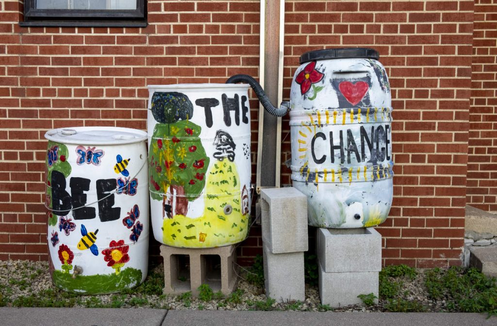 Three white barrels painted with bees, trees, and flowers with the words BEE THE CHANGE against a red brick wall
