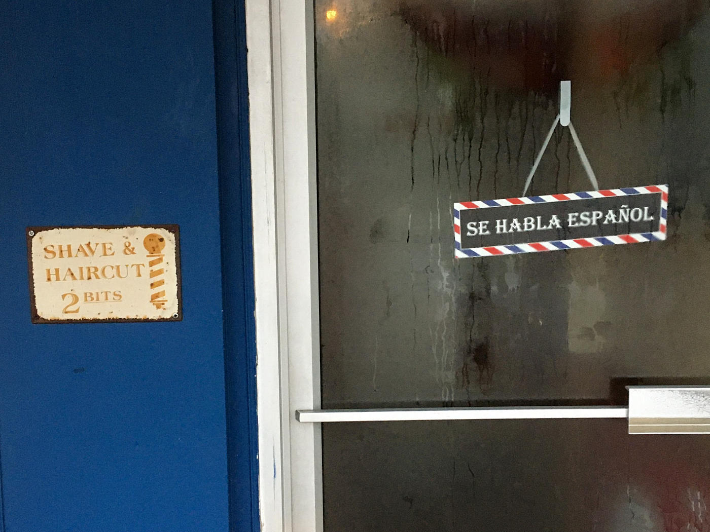 Faded sign on blue wall reading "SHAVE AND HAIRCUT 2 BITS" next to a glass door with a sign reading "Se Habla Español,"