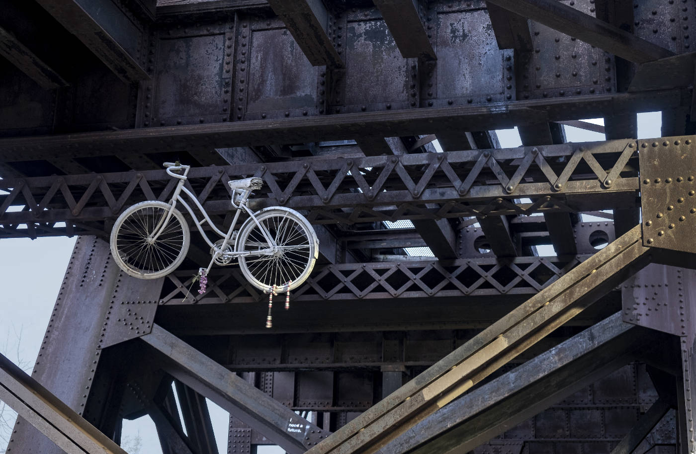 white coaster bicycle hanging under a dark gray metal superstructure under a bridge
