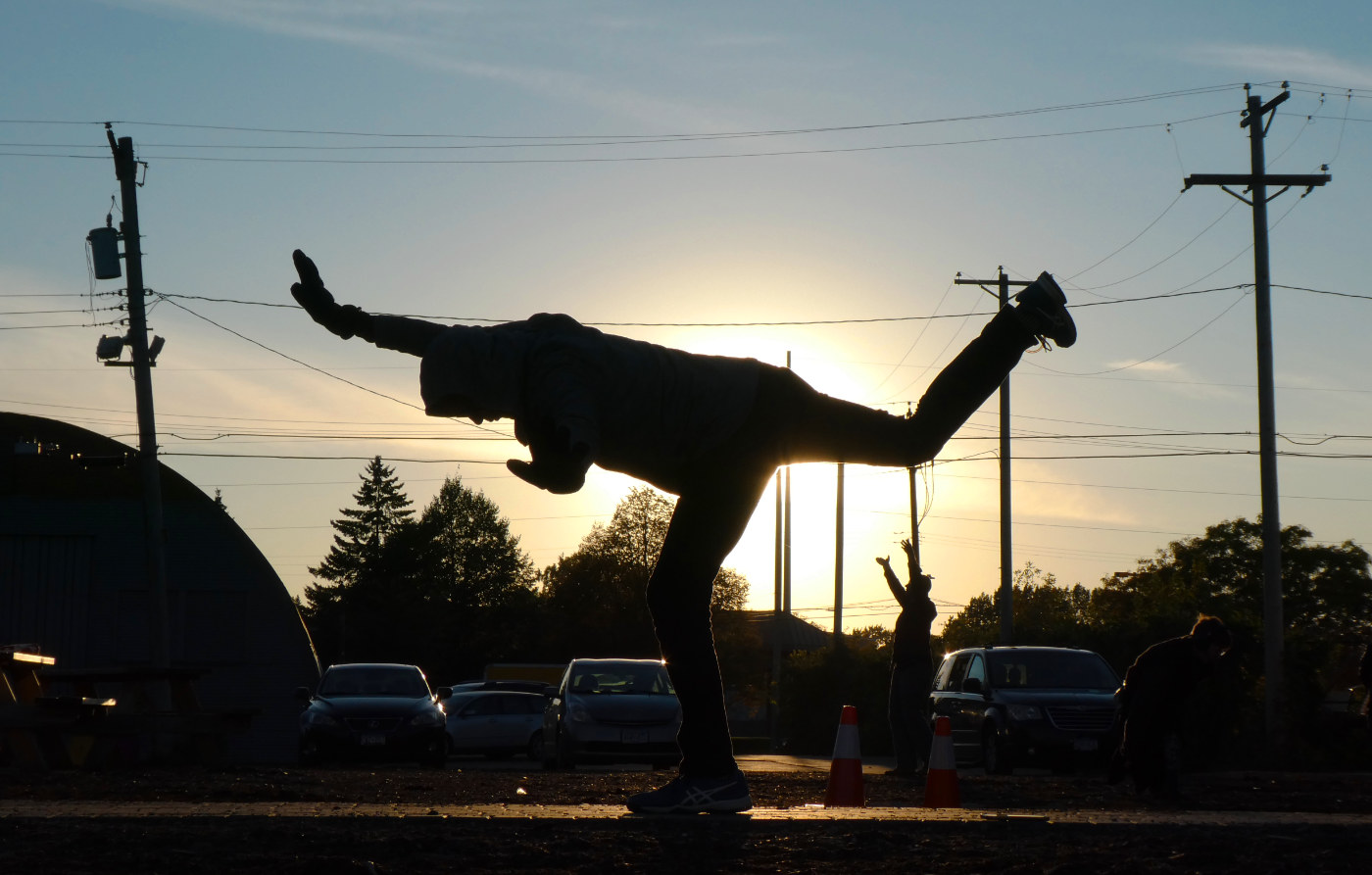 silhouette of figure standing on a single leg with leg outstretched backwards and arm outstretched forward against a setting sun framed by two telephone posts and lines