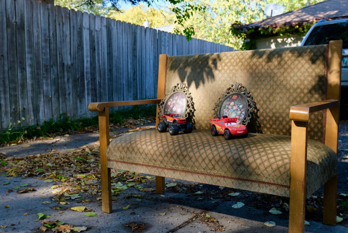Aged brown upholstered settee with a pair of small floral prints and pair of red toy trucks in a driveway