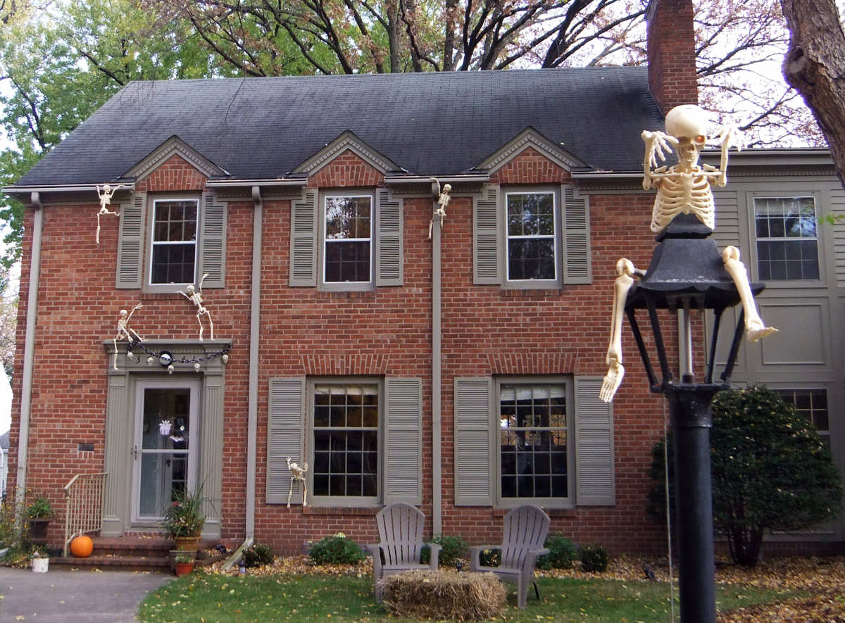 A skeleton atop a lamppost in front of a two-story brick house with skeletons climbing up the wall