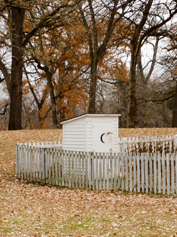 Small white shack with a half moon shape cut out of door behind a white picket fence in a field of brown leaves with trees in background