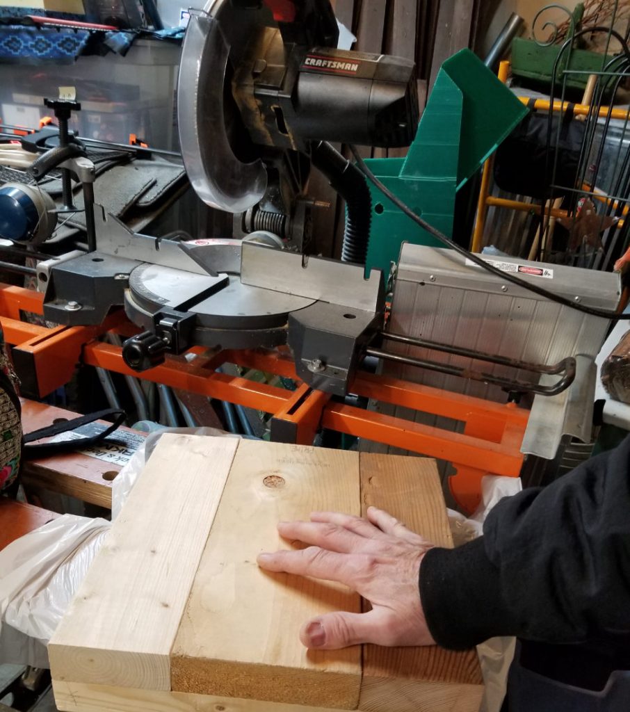 Human hand on wood pieces next to a large steel circular saw 