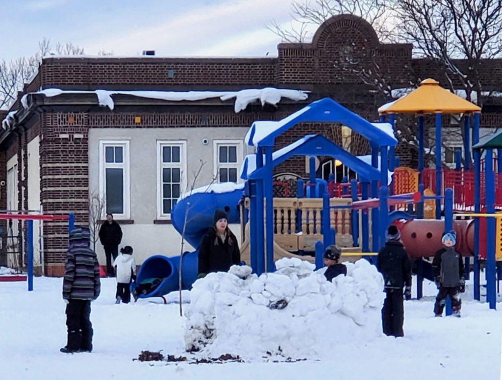 children in a snow-covered playground with one small boy behind a snow wall