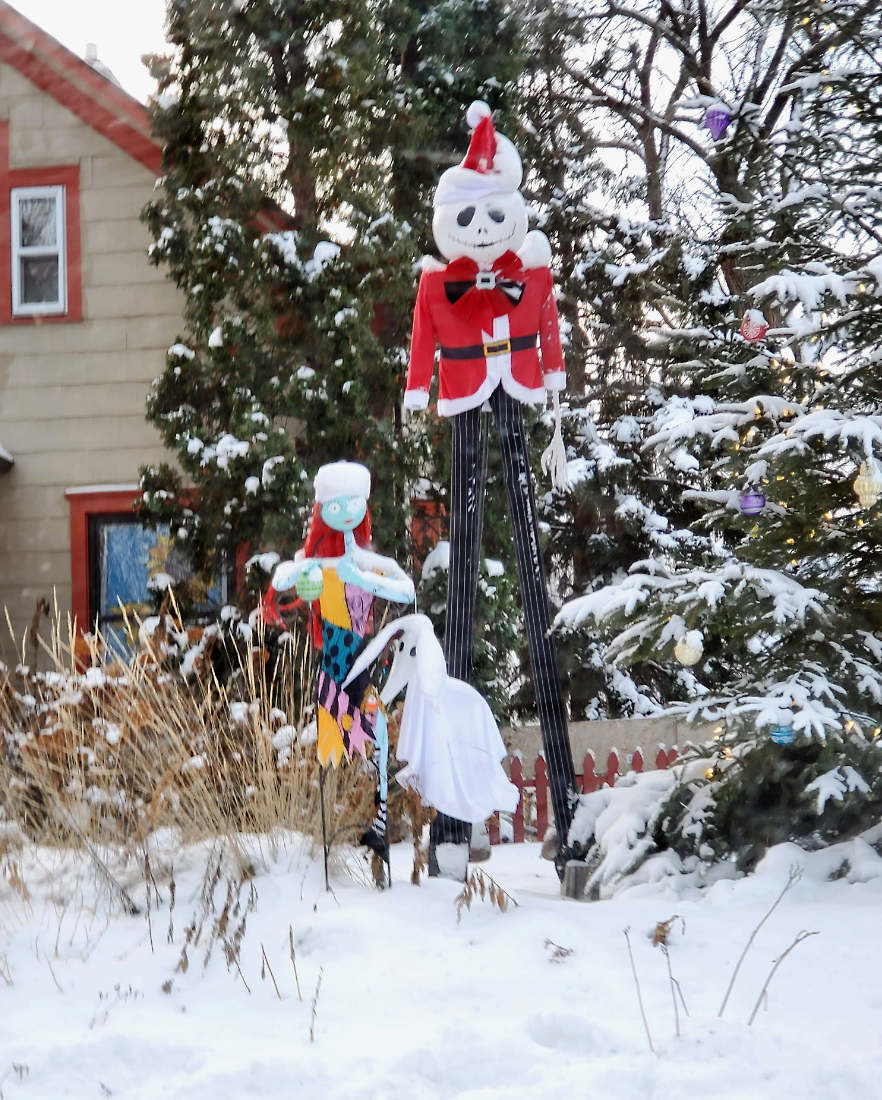 Tall scarecrow-like statue with a red and white Santa coat and hat with some other figures in a snowy scene outside of a house