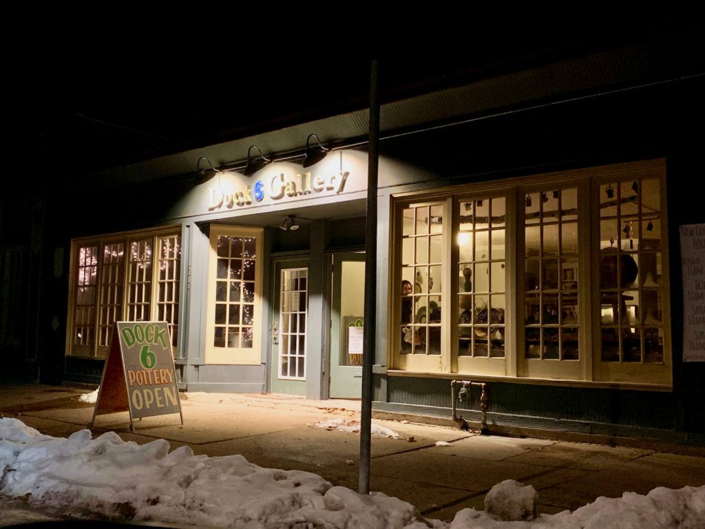 night scene of storefront lit by three overhanding lights with a sandwich sign on a snow-shoveled sidewalk