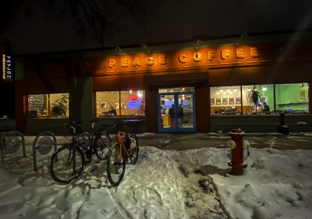 Night scene with snowy street, with parked bicycles, and brightly colored storefront windows with yellow-orange overhead sign reading "peace coffee"