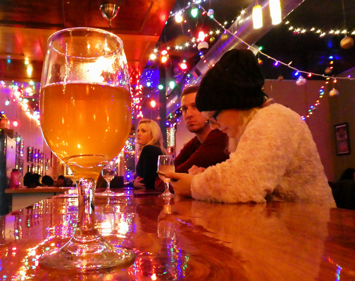 closeup of a wineglass half-filled with golden liquid on a bartop with people leaning on it in background with many colored lights