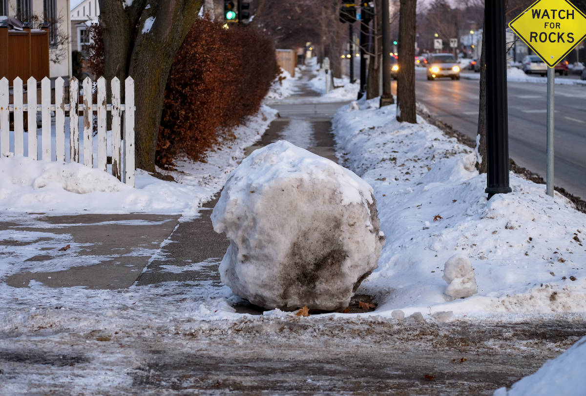 Very large snowball on a sidewalk in a snowy street scene with cars & lights in background