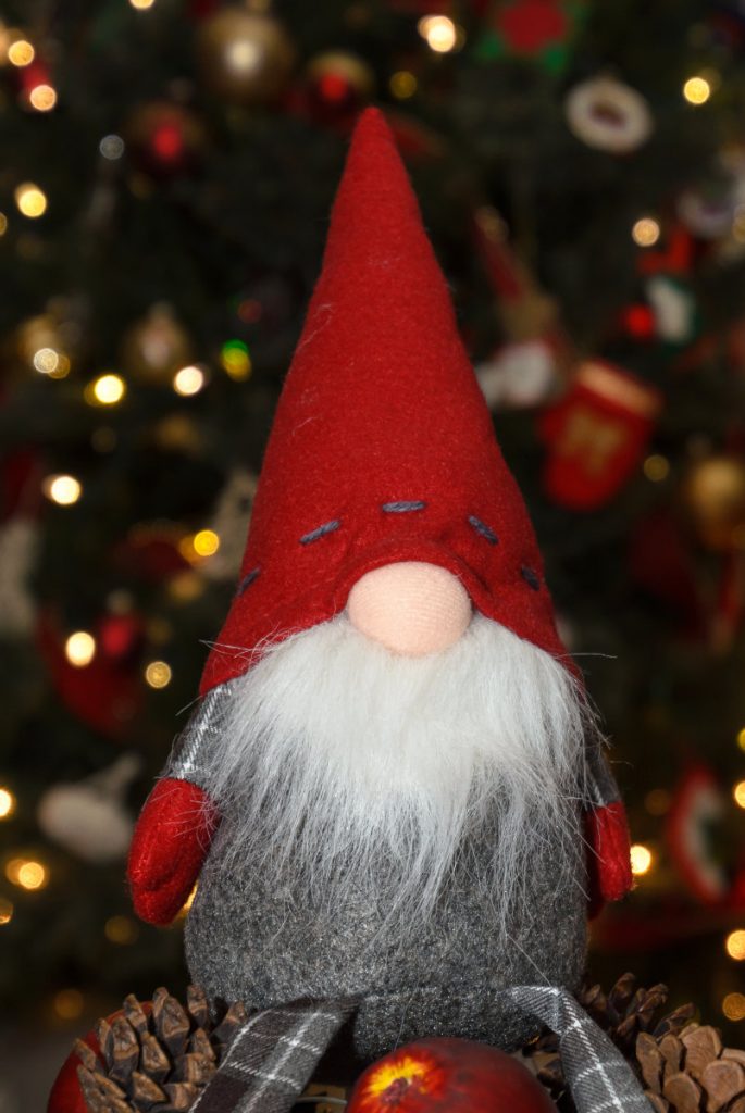 Elfish doll with conical red hat down over eyes, oval pink nose, and grayish-white beard with blurred christmas tree lights in background