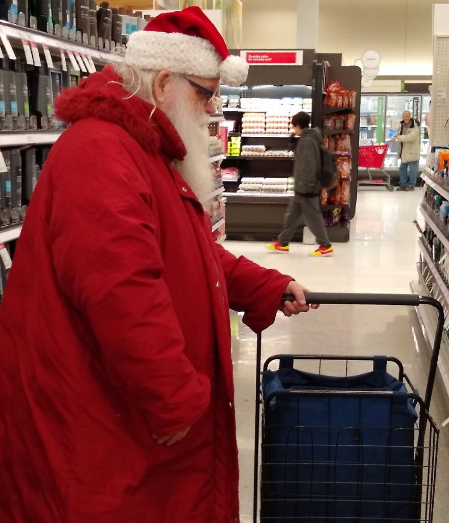 old man with gray-white beard in red coat and santa hat in a store aisle with cart