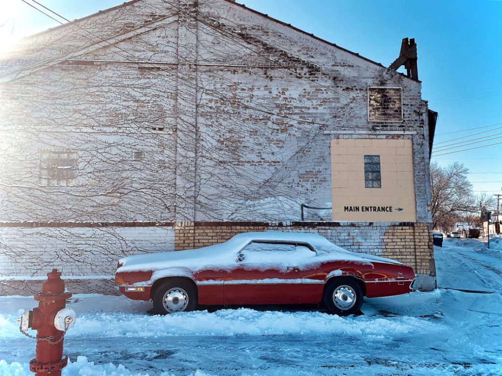 old classic red car covered in snow next to gray warehouse building