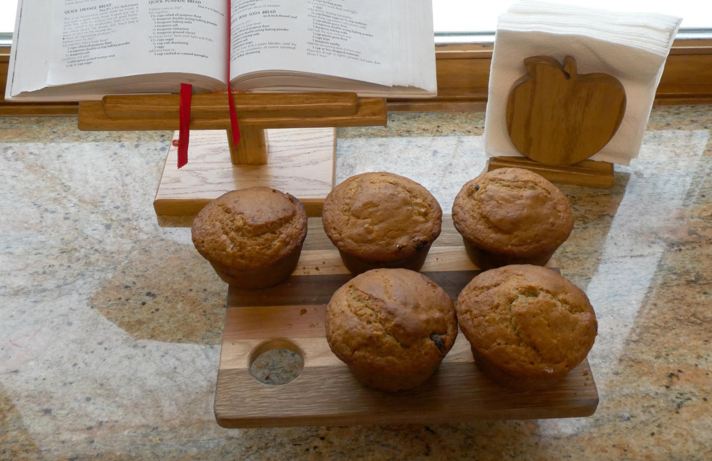caramel brown muffins on a breadboard on a granite counter withwooden cookbook stand and apple-shaped wooden napkin holder.