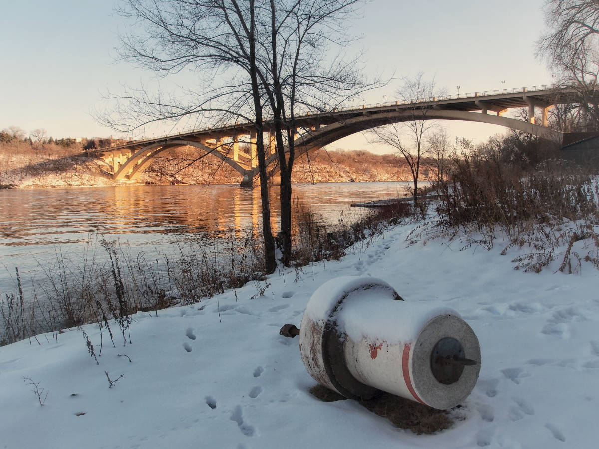 riverside scene with snow-covered bank with buoy on it's side, with sunlit bridge over river