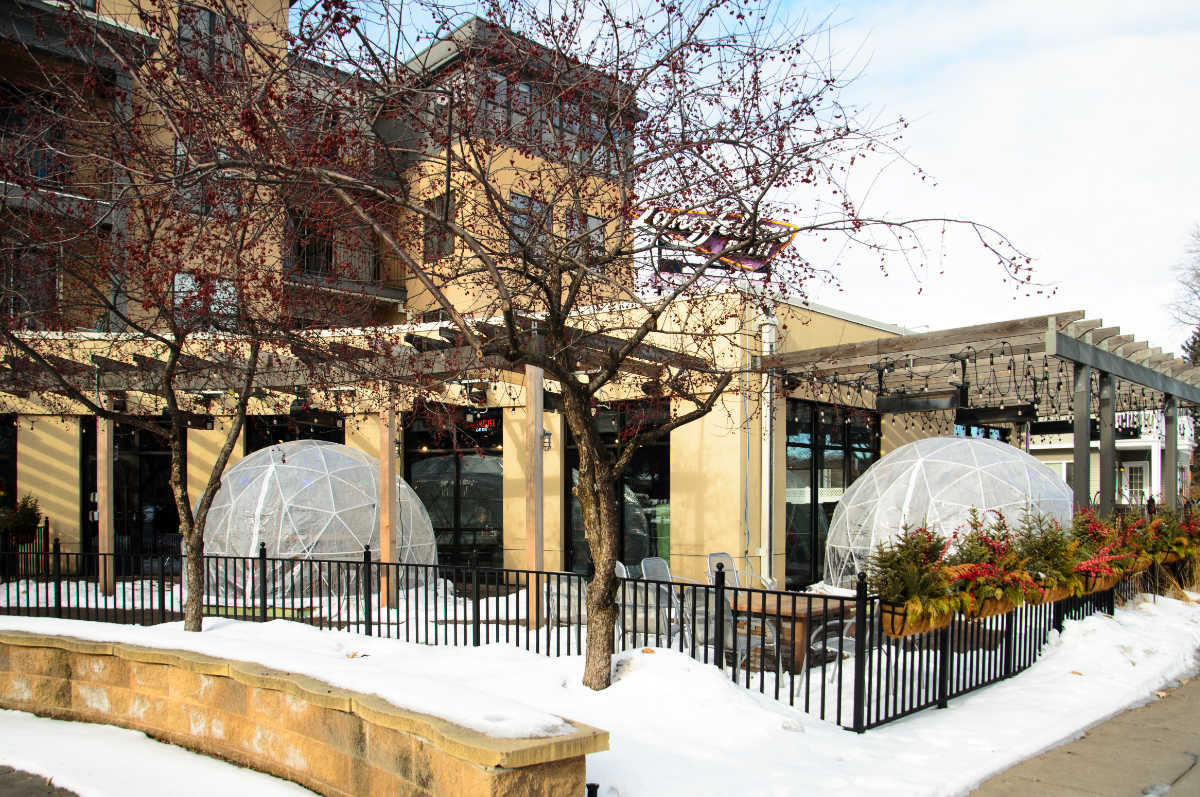 snow-covered patio outside yellow stone building with yellow stone bench and two plastic geodesic domes behind a black iron fence