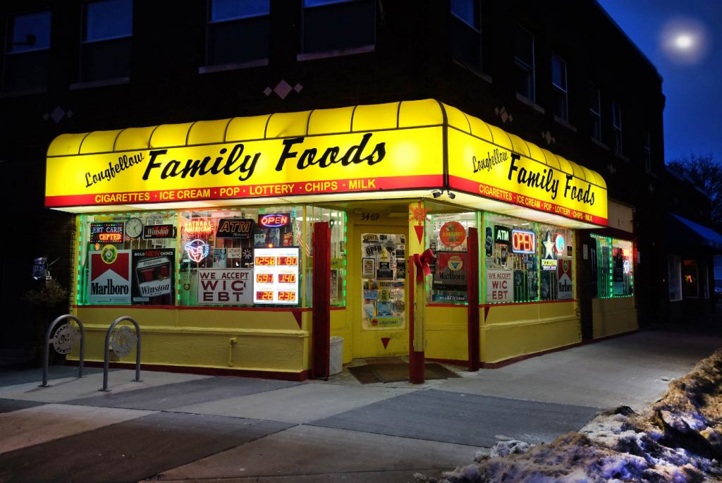 Corner convenience store at night with lit yellow awning and Longfellow Family Foods across both sides