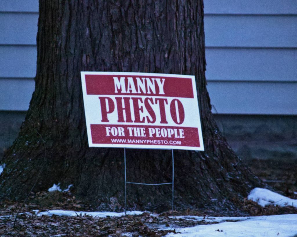 Red and white lawn sign leaning on a tree trunk reading "MANNY PHESTO FOR THE PEOPLE"