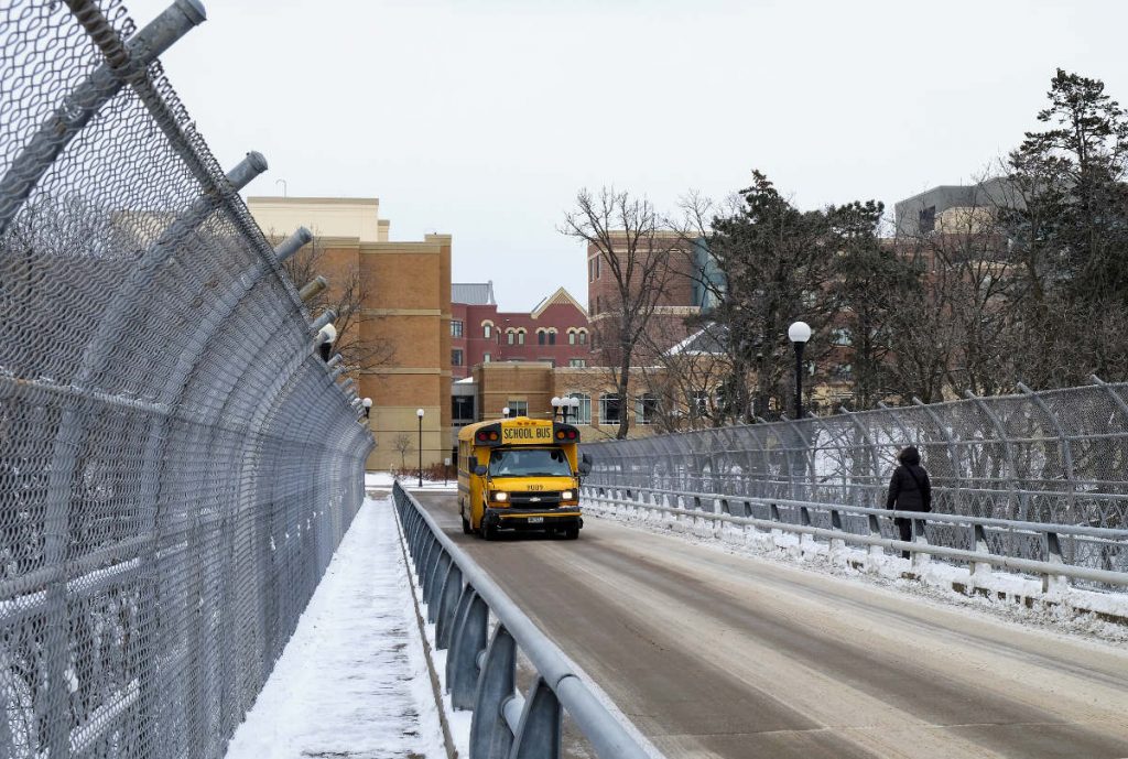 yellow school bus approaching on bridge road with sidewalk and fencing on right side with buildings in background