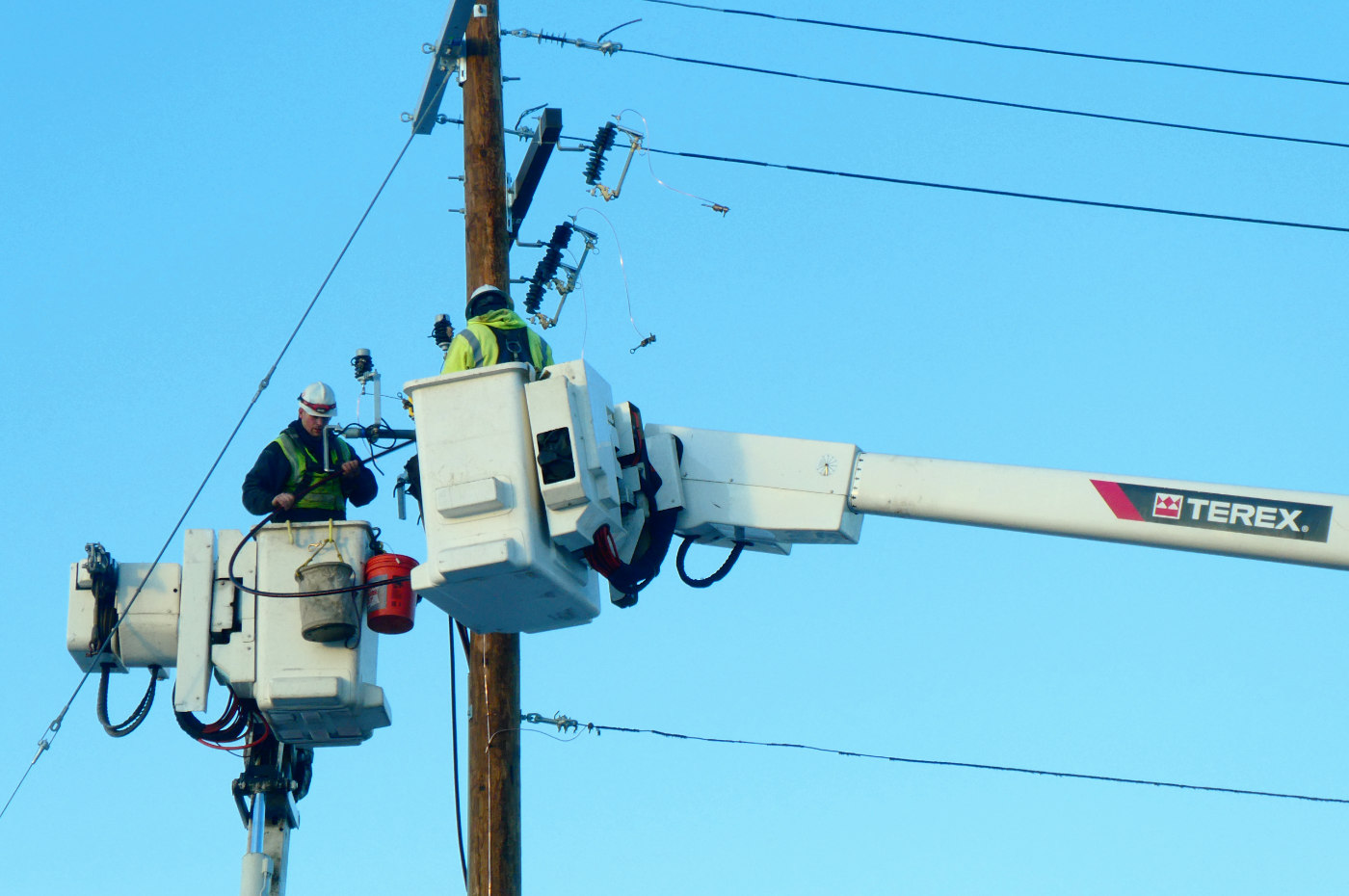 two men in white cherry pickers working on telephone pole wiring against a bright blue sky