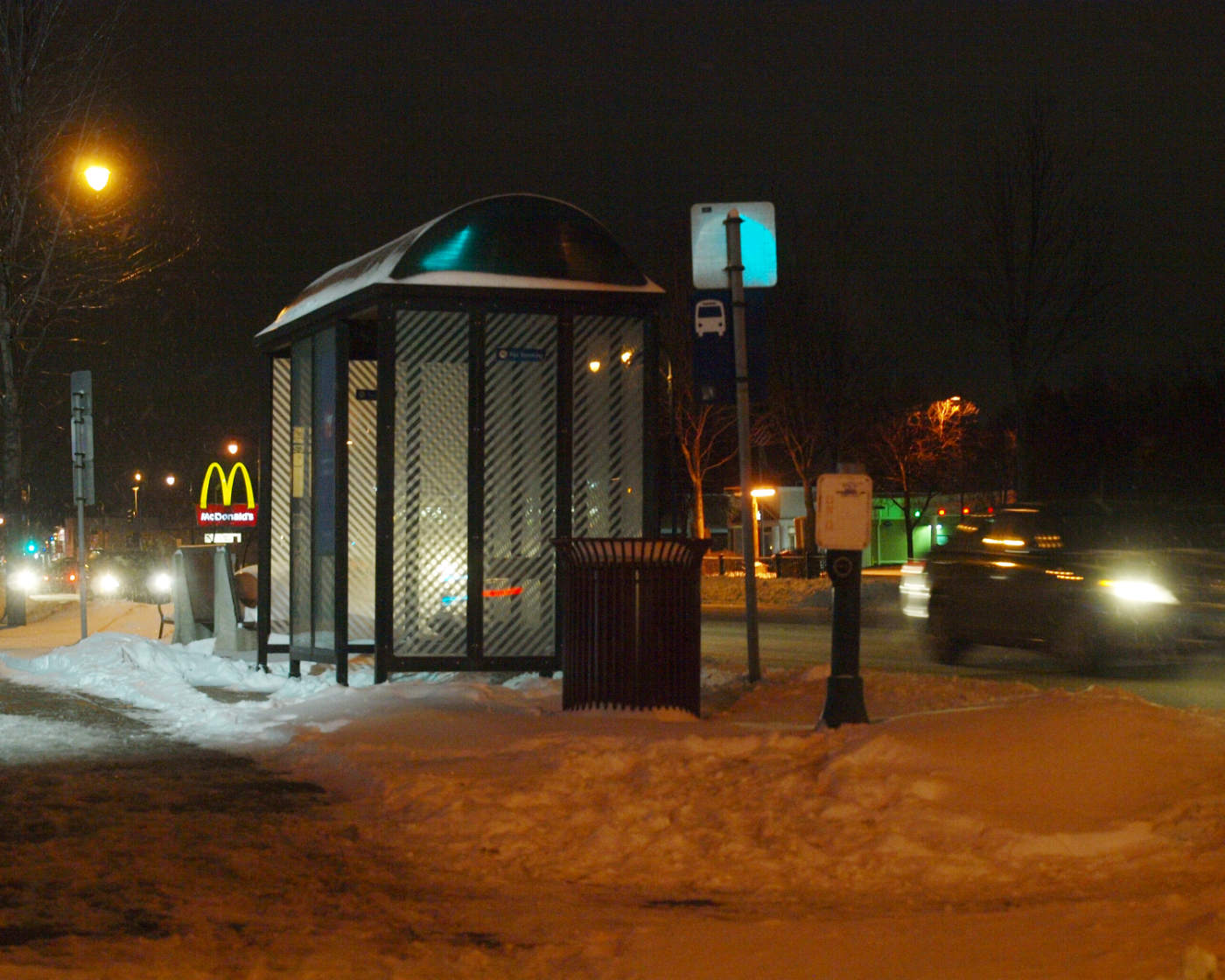 Snowy night scene of bus stop lit by streetlights and car lights with retail signs in background