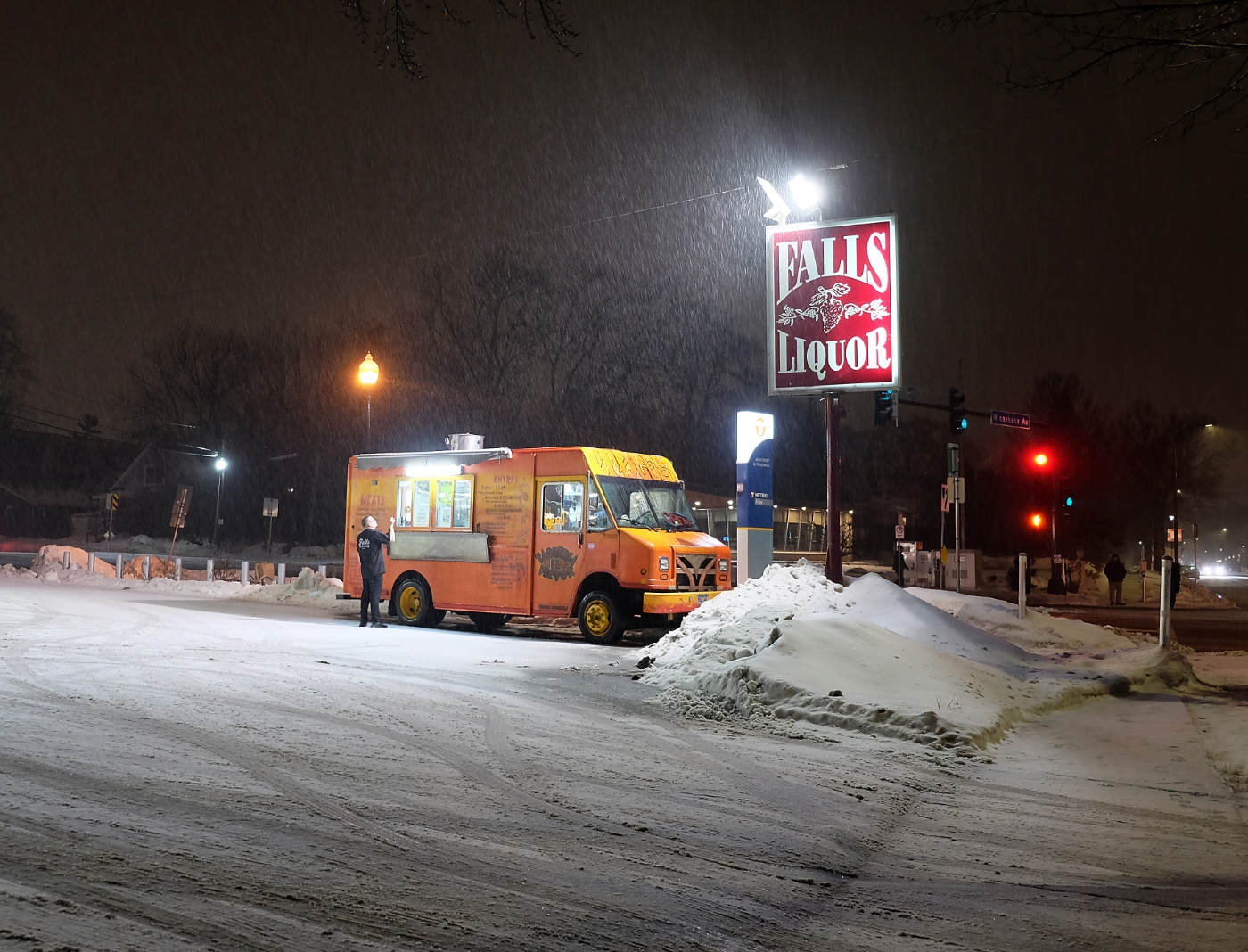 Snowy parking lot at night with an orange food truck under a street light with a person ordering at the truck window, and a red and white FALLS LIQUOR sign next to it