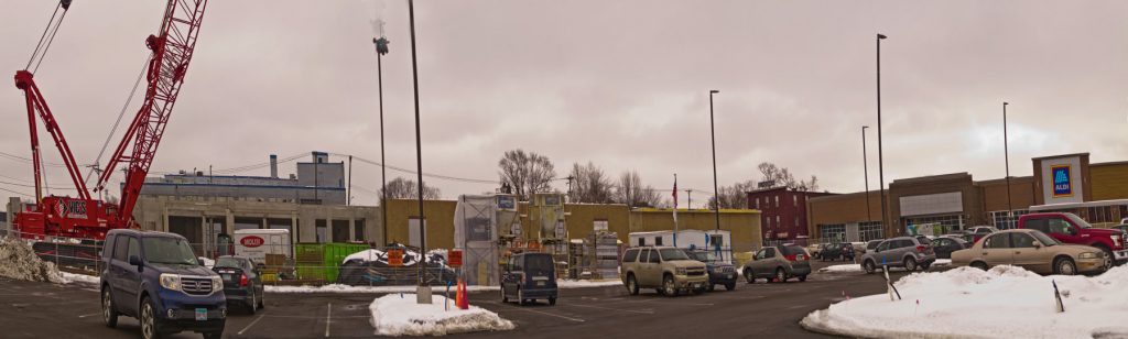 panoramic view of construction site with cars in a parking lot, low buildings, and a tall red construction crane