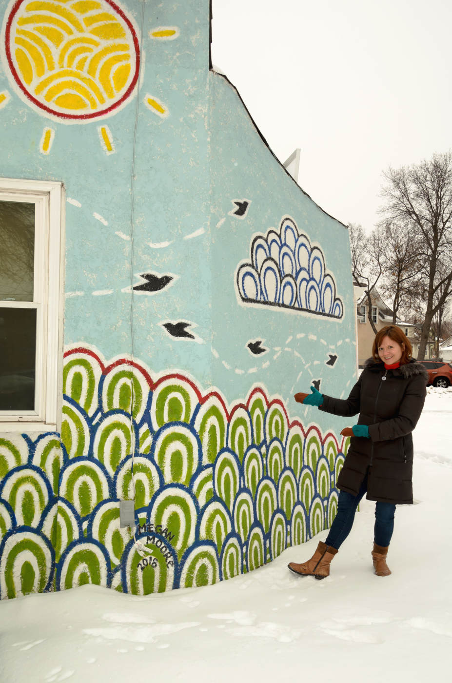woman standing next to a single story building with a mural of an abstract landscape with a field of green globe shapes with bird silhouettes flying in a blue sky under a yellow sun