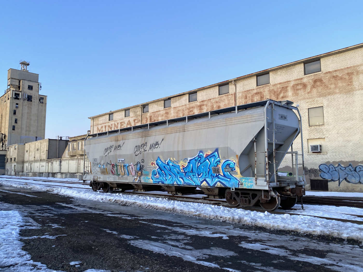 railroad car with some color graffiti in front of a long yellow stone building labeled MINEAPPLIS SEED COMPANY with a tall grain elevator against a blue sky