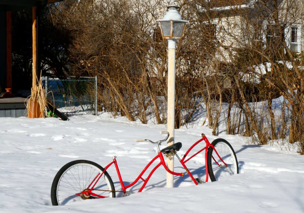 red tandem bicycle missing one seat and one handlebars leaning against a lamppost in deep snow with brush and houses in the background on a sunny day
