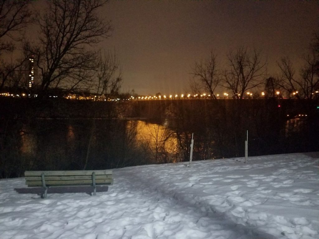 snowy night scene of park bench facing bridge lights with reflections on the water.