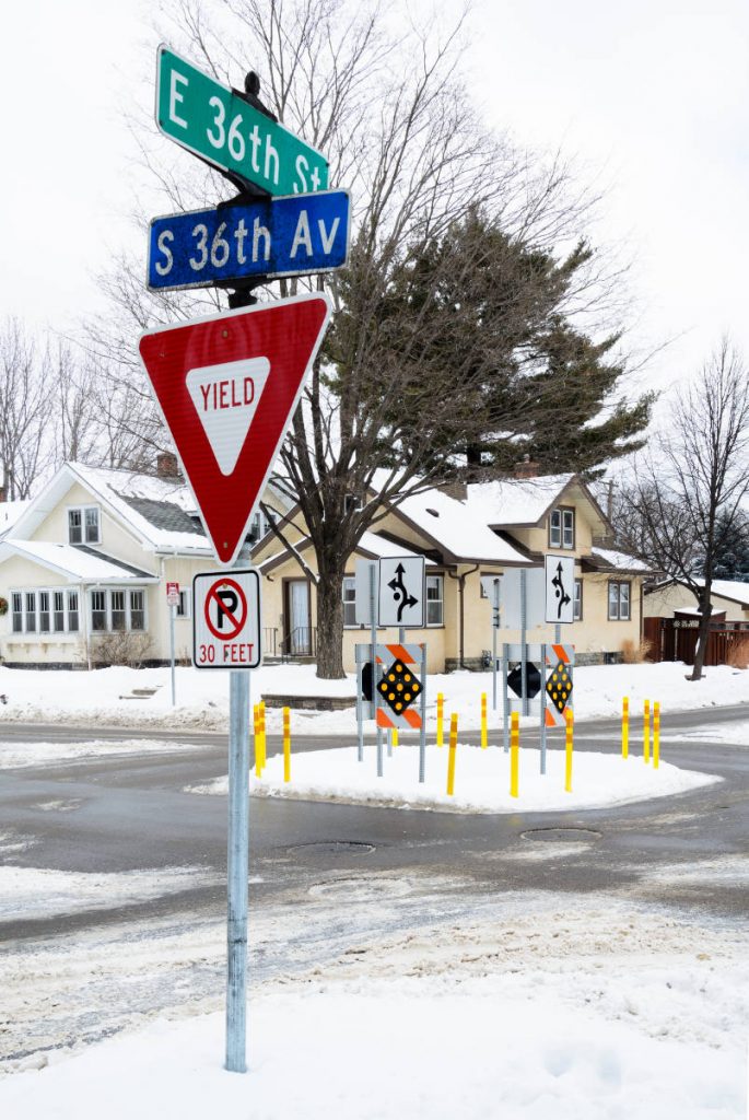 Red and white yield traffic sign with S 36th Av and E 36 St street signs with an intersection in the background with a traffic circle of yellow posts and diamond-shaped reflective signs