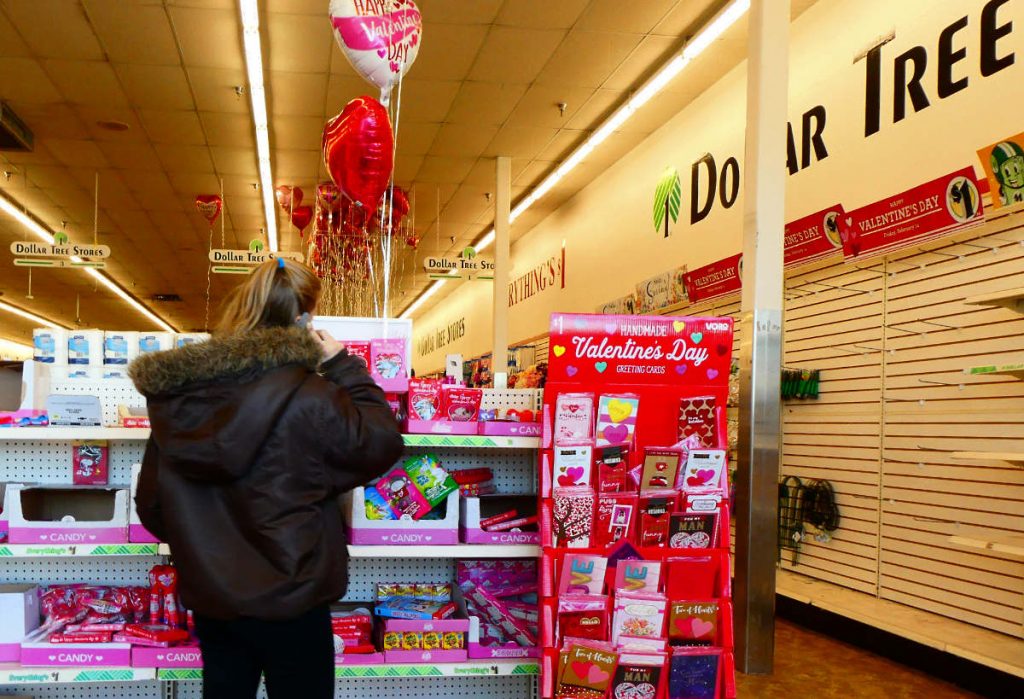 back of woman in brown leather winter coat standing in aisle of Valentine's Day cards and gifts with helium balloons overhead and Dollar Tree signage on the wall