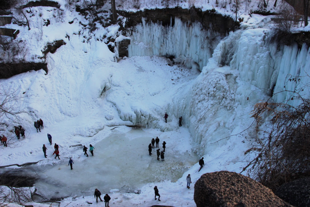 High overview of frozen waterfall, pond, and surrounding cliffs with people milling around at bottom