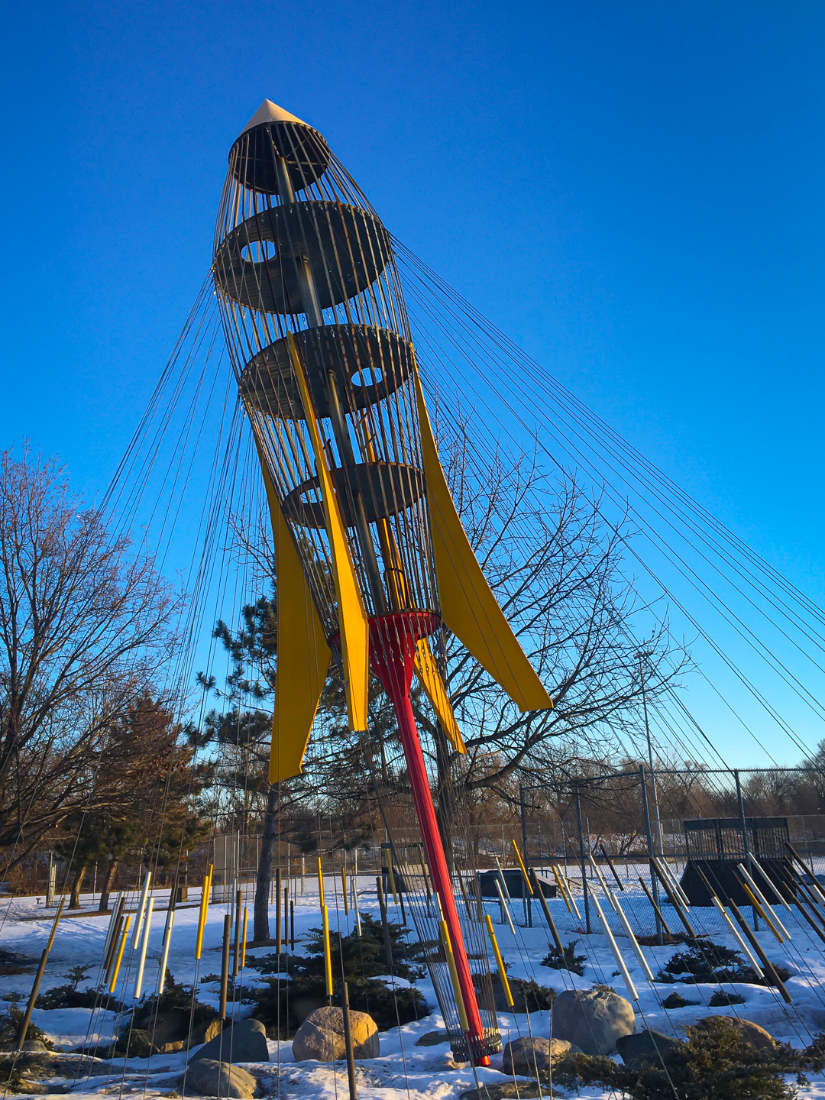 Tall see-through rocket-shaped metal sculpture with yellow fins on a red pole with guy-wires against a blue sky.