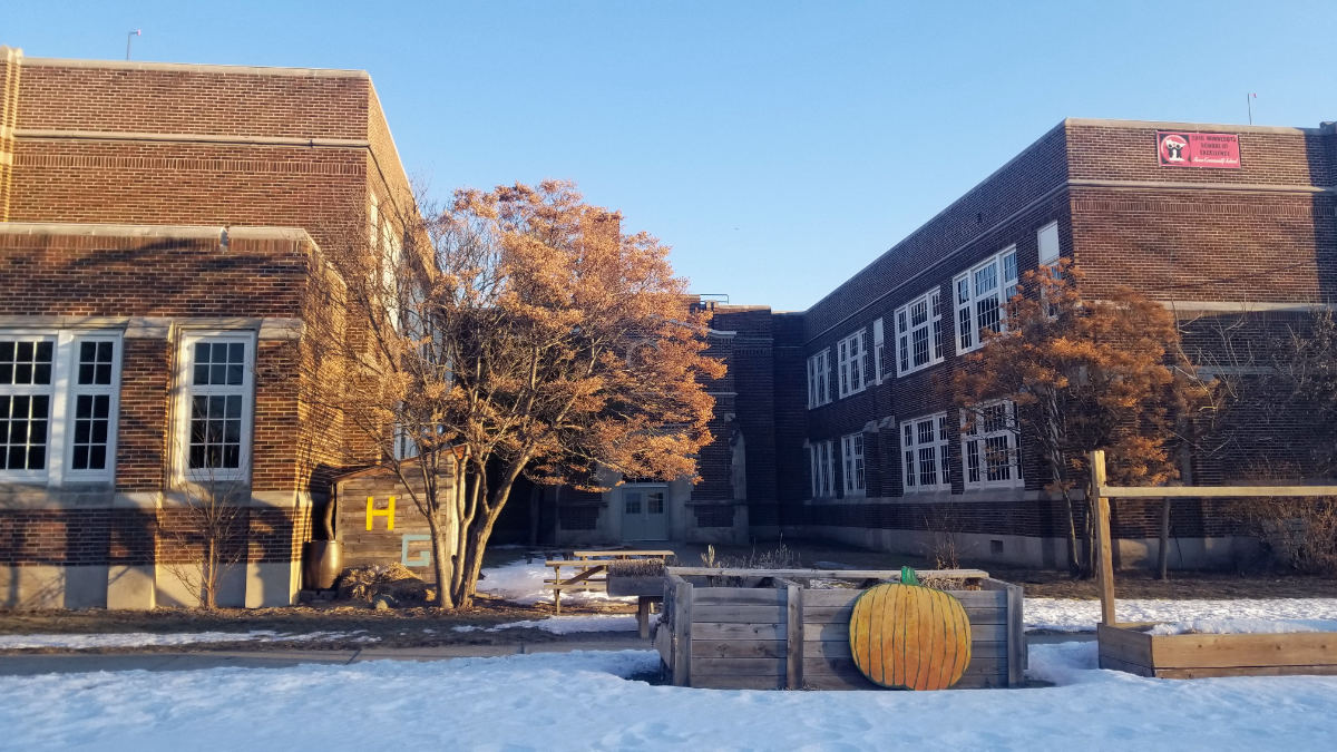 Two-story orange brick school building fronted by a wooden planter box with a pumpkin placard on it in a sunny winter scene