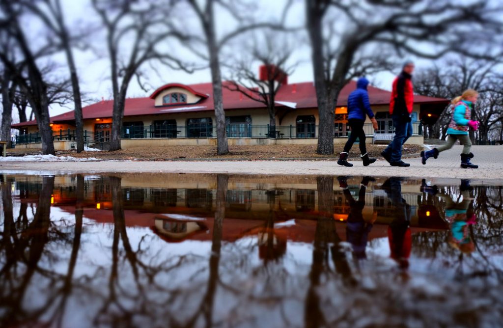A man and two children bundled in winter clothes reflected in a large puddle of water with a park building in the background