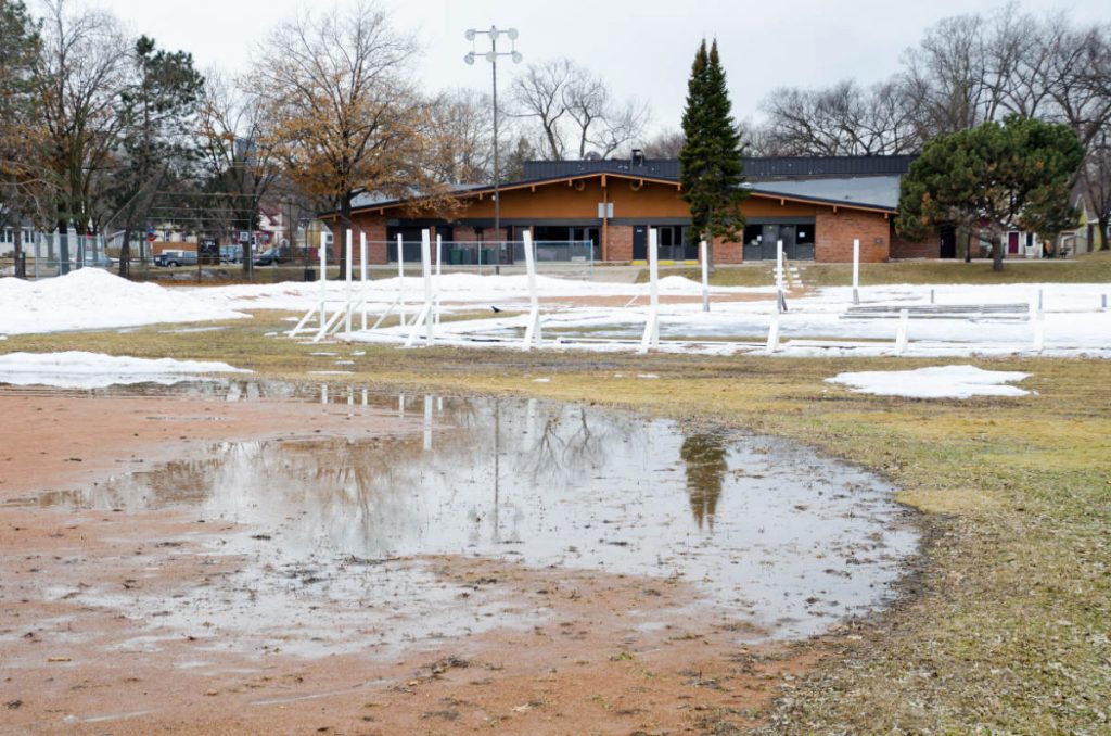 reflective puddle of water on dirt next to brown grass and low snow mounds with a park building in the background