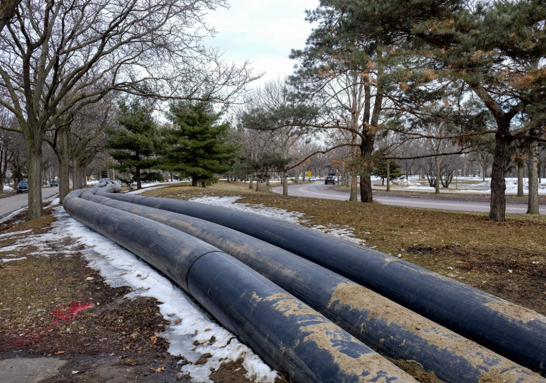 three long blue large snaking pipes run from foreground to background over grass along a parkway