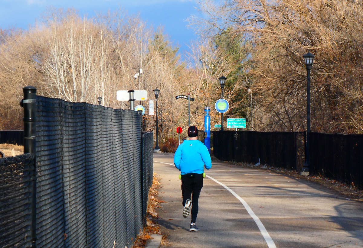 A runner in sky blue jacket and black clothes seen from behind on a cement trail divided with a white line with black fencing and signage beneath a bright blue sky over bare trees