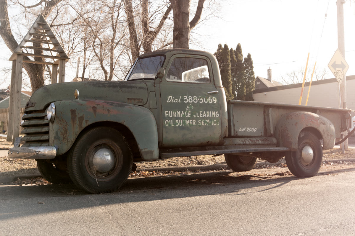 an antique gray truck parked on the street, with commercial painting on the door reading: Dial 3883069, for Furnace Cleaning , Oil Burner Service