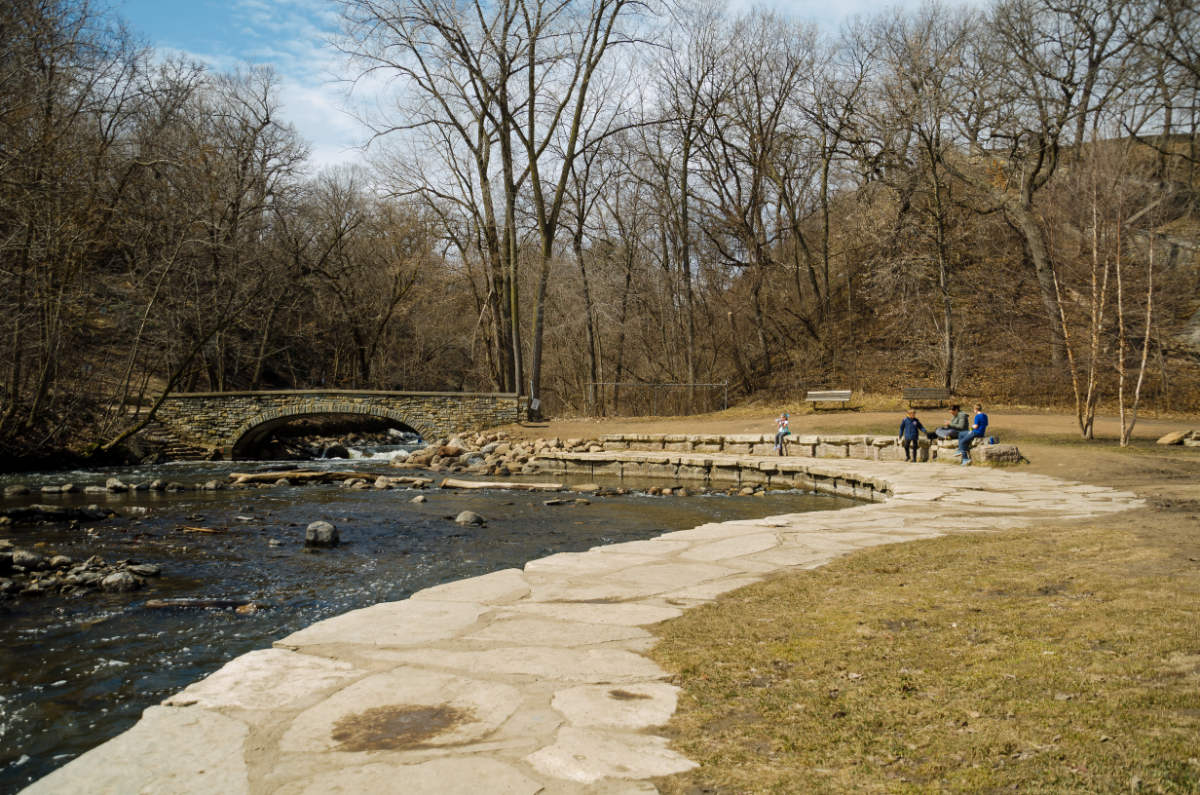 stone walkway along creek with people sitting on a bench and a stone bridge over creek in background