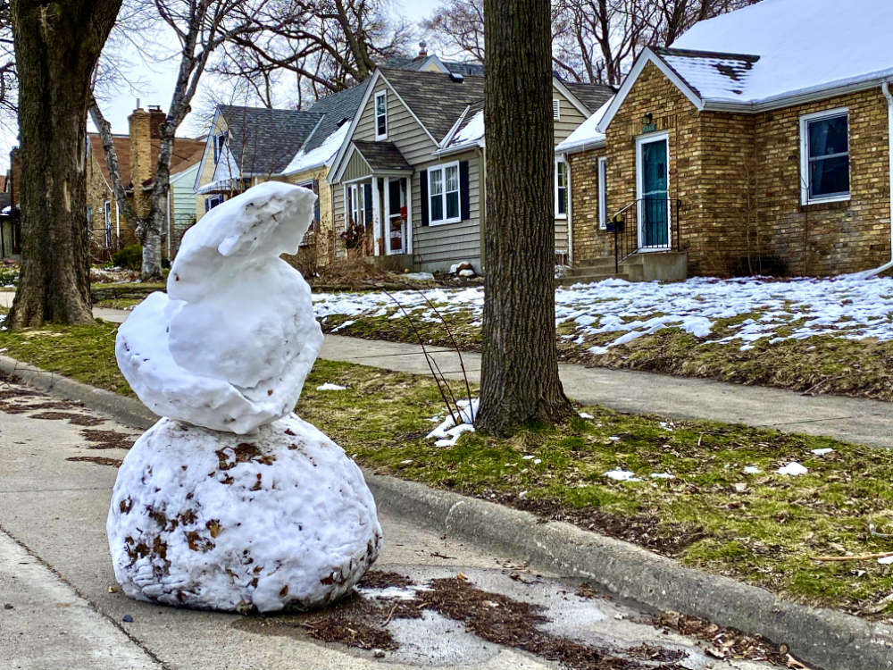 large tilting snowman on a city street next to a grassy boulevard with somewhat snowy lawns and houses in the background