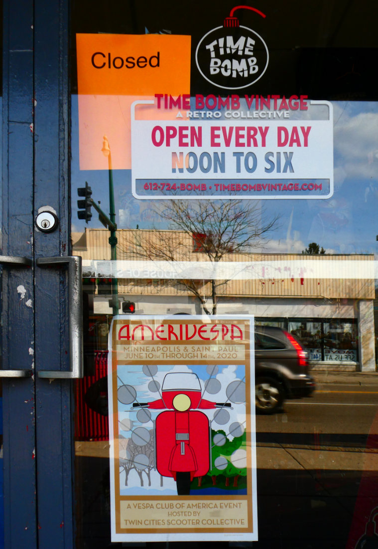 Signage on a glass storefront door: Open Every Day Noon to Six with a placard reading Closed above it, a colorful poster of a scooter for AMERIVESPA