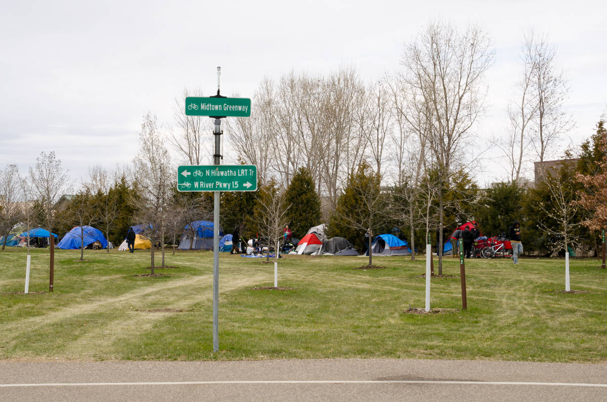 Green space behind paved path with green direction signs with a row of tents against a line of evergreen trees in the background