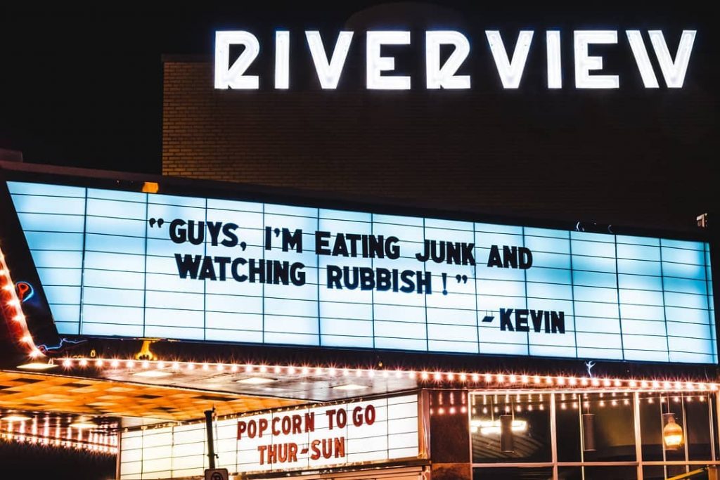 Theater marquee at night reading "Guys, I'm eating junk and watching Rubbish! -Kevin" under RIVERVIEW white lights