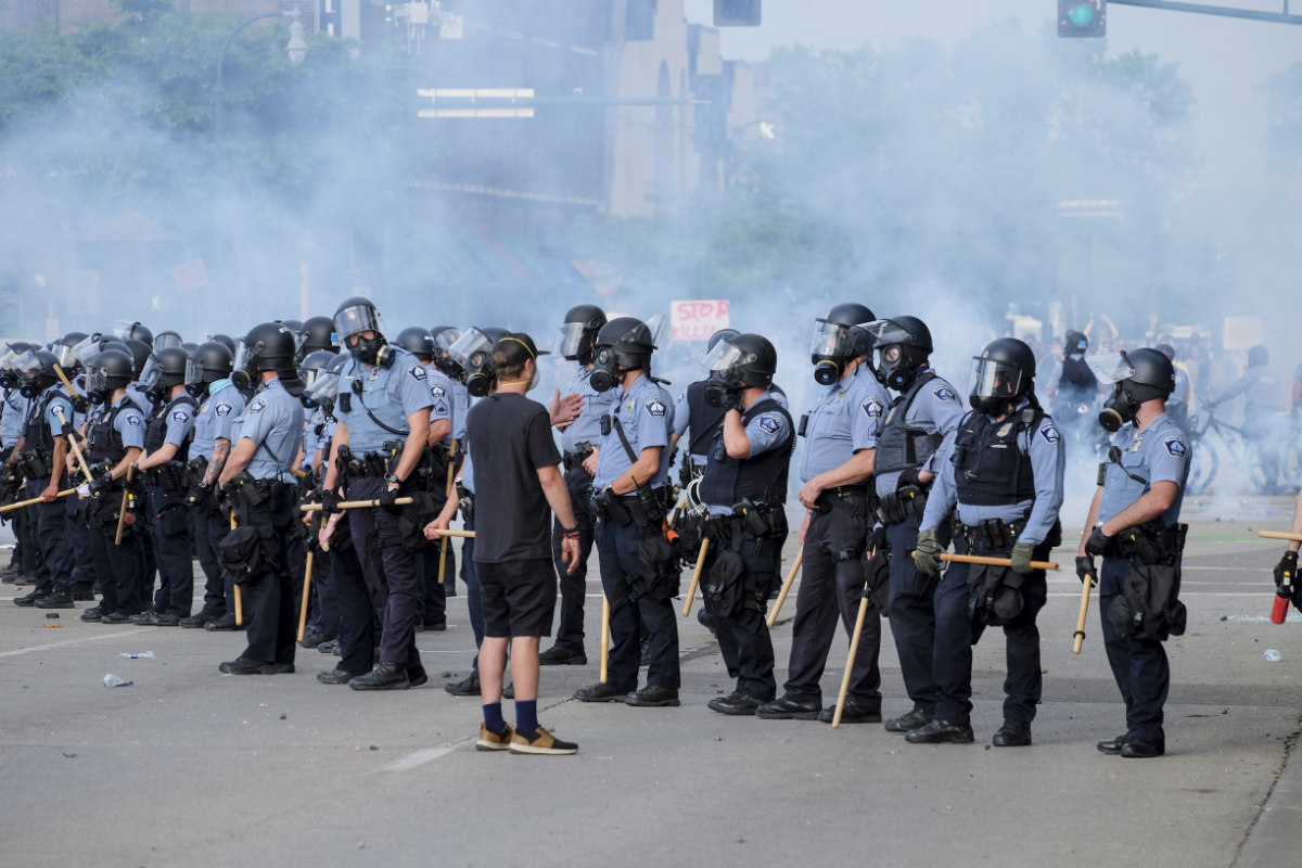 A long line of over a dozen police officers in blue and black uniforms with riot gear facing a single young man in black shorts and t-shirt and a white face mask addressing the police with one hand up gesticulating as though talking