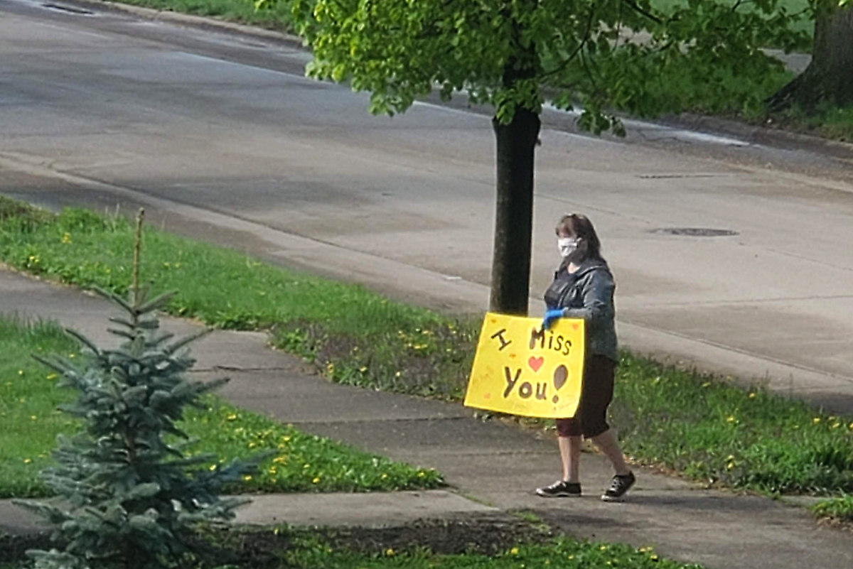 Woman wearing a medical mask standing on a sidewalk holding a sign that says "I Miss You" with a red heart in the middle.